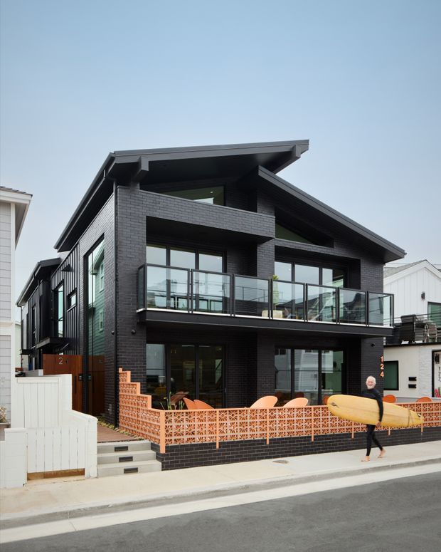 Street-facing exterior photograph of a contemporary two-storey black brick house with a prominent asymmetrical gable roof, large glazed openings, a glass-balustraded upper balcony, and a patterned terracotta breeze-block front fence. A narrow entry stair and dark brick boundary wall lead from the sidewalk to the home.