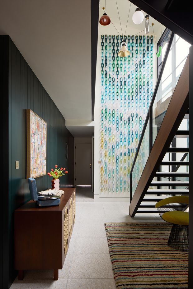 A contemporary residential entry hall with a dramatic open stairway, dark vertical wall panelling, pale tiled flooring, and a tall glazed stair void. A colourful chain-link installation forms a striking feature wall, while pendant lights, a timber console, artwork, and a patterned rug add a playful, eclectic character.
