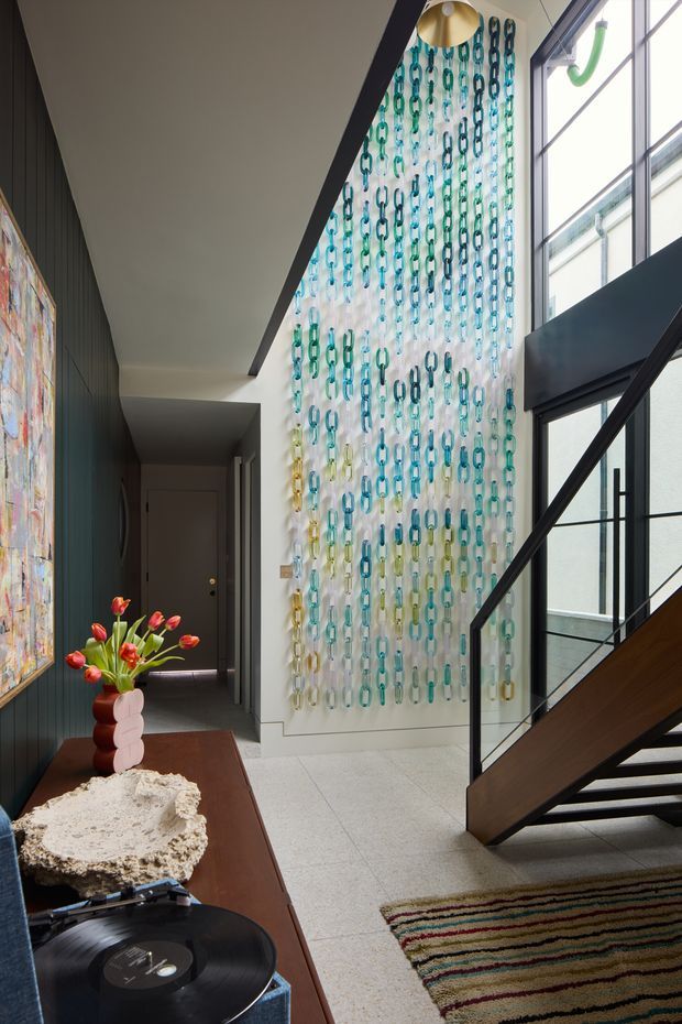 A contemporary residential entry hall with a dramatic stairway and tall glazed opening. The space features dark vertical wall panelling, light tiled flooring, black-framed glazing and stair balustrades, a colourful hanging chain installation across a double-height wall, and a mid-century-inspired timber console with artwork, flowers and a record player. A striped rug adds colour at the base of the stairs.