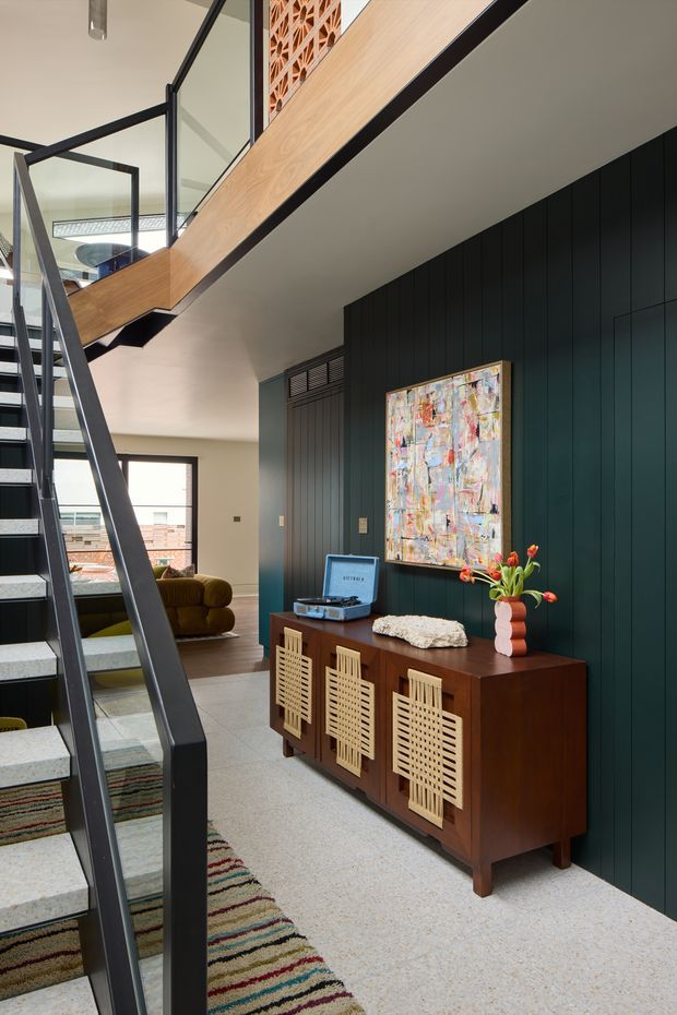 A stylish residential stairway and landing with a dark metal open-stringer staircase, pale stone or concrete-look treads, and a timber-clad stair balustrade. The landing features dark vertical wall panelling, a mid-century wooden sideboard with decorative woven-front doors, a turntable, artwork, and a sculptural vase. A living area with a mustard sofa is visible beyond, while a patterned runner adds colour to the floor. The interior has a contemporary design with mid-century furnishings and strong contrasting timber, black, and neutral tones.