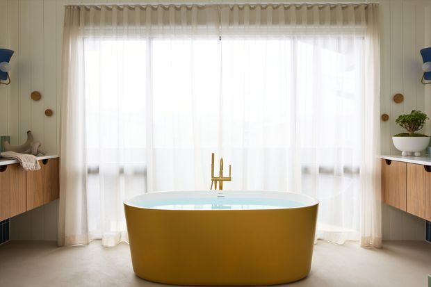 Bright, symmetrical bathroom featuring a freestanding mustard-gold bathtub centered in front of full-height sheer curtains and a large window. Pale vertical wall panelling, warm neutral flooring, floating timber vanity cabinetry on both sides, wall-mounted brass-toned fixtures, and minimal decorative objects create a refined mid-century-inspired interior.