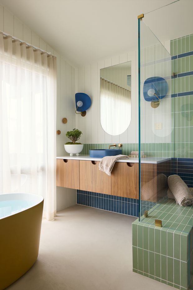 Bright, playful contemporary bathroom featuring a freestanding bathtub, floating timber vanity with integrated basin, large rounded mirror, and a glass-enclosed tiled shower. The room combines pale vertical wall panelling with green and navy blue grid tiles, warm timber cabinetry, rounded fixtures, and sheer curtains. A built-in tiled bench or ledge is visible within the shower area.