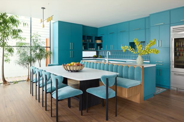 A bright, spacious open-plan kitchen and dining area with bold teal-blue full-height cabinetry, integrated appliances, a large pale stone island, and a matching curved upholstered banquette. A long dining table with turquoise chairs occupies the foreground, while tall glazing and indoor greenery bring in abundant natural light. Warm timber flooring contrasts with the cool cabinetry and upholstery.