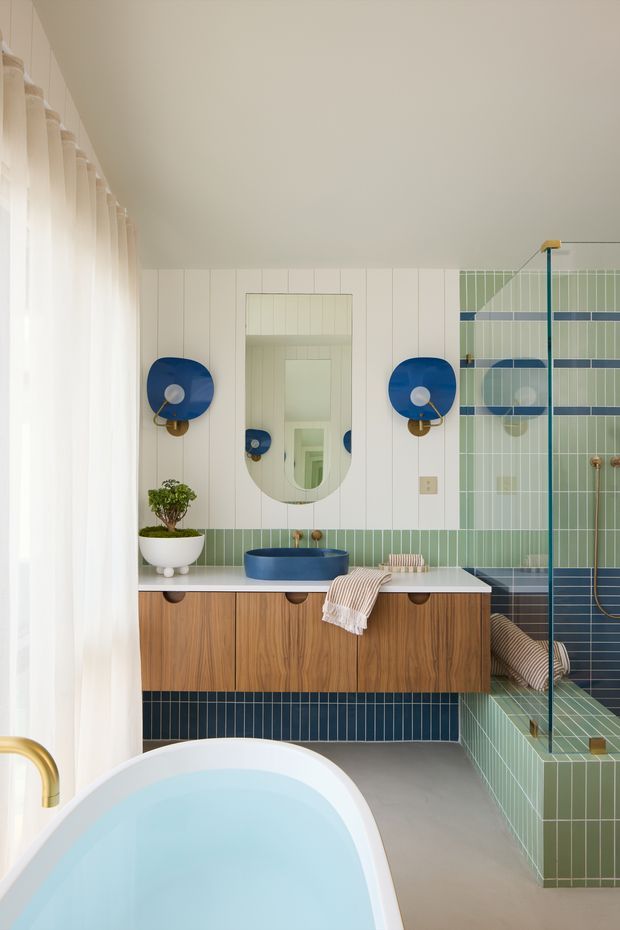 A bright, carefully styled bathroom with a freestanding white bathtub in the foreground, a floating timber vanity with a deep blue vessel basin, and a large mirrored medicine cabinet. The room features pale green vertical wall and shower tiles, dark blue horizontal tile accents, blue circular wall sconces, brass fittings, white vertical wall panelling, and a frameless glass shower enclosure. Sheer curtains provide soft filtered daylight along the left side.