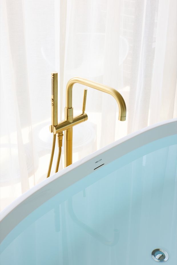 Close-up product-style photograph of a freestanding bathtub with a tall brushed gold/brass tap and handheld shower fixture. The tub has a pale blue interior and white exterior edge, set against softly diffused white sheer curtains.