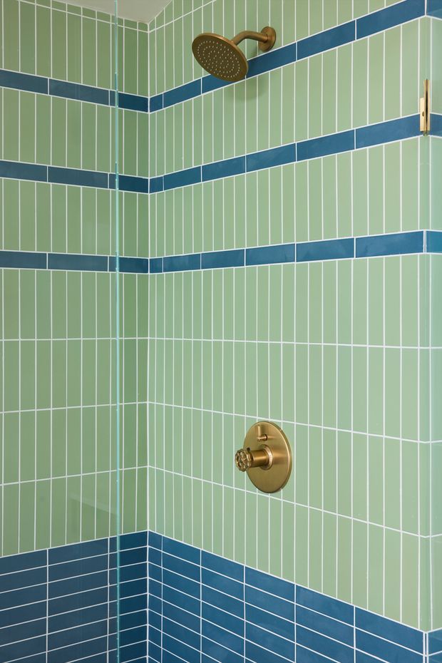 Close-up view of a tiled shower enclosure with pale mint-green vertical rectangular wall tiles, repeated dark blue horizontal accent bands, and dark blue lower wall tiles. The shower hardware, including a round showerhead and control, has a warm brass finish. A frameless glass shower panel is visible at the corner.