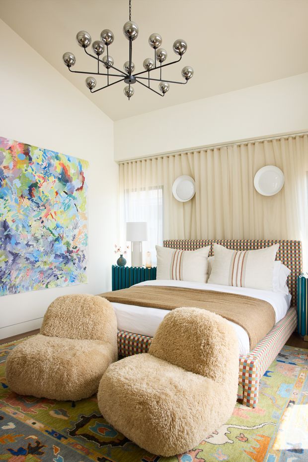 A bright, highly styled bedroom featuring a bed with a colourful geometric upholstered frame and headboard, layered neutral bedding, two plush rounded boucle lounge chairs, a large vibrant abstract artwork, patterned area rug, teal ribbed radiator cover or console, pleated cream curtains, and a sculptural multi-arm globe chandelier. The room combines playful colour and pattern with soft, tactile furnishings and contemporary statement lighting.