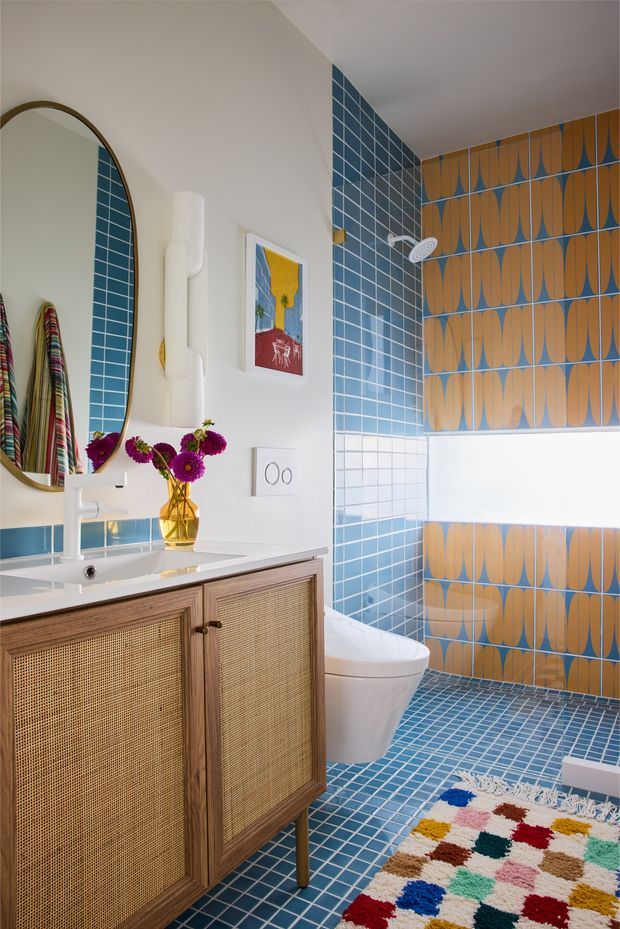 A colourful, retro-inspired bathroom with a blue tiled floor and shower surround, featuring patterned blue-and-mustard wall tiles. The room includes a white wall-mounted basin with a timber vanity clad in woven cane panels, a large rounded-edge mirror, white plumbing fixtures, a framed artwork, and a multicoloured woven bath mat. The space is bright and playful with strong geometric patterning and warm timber accents.