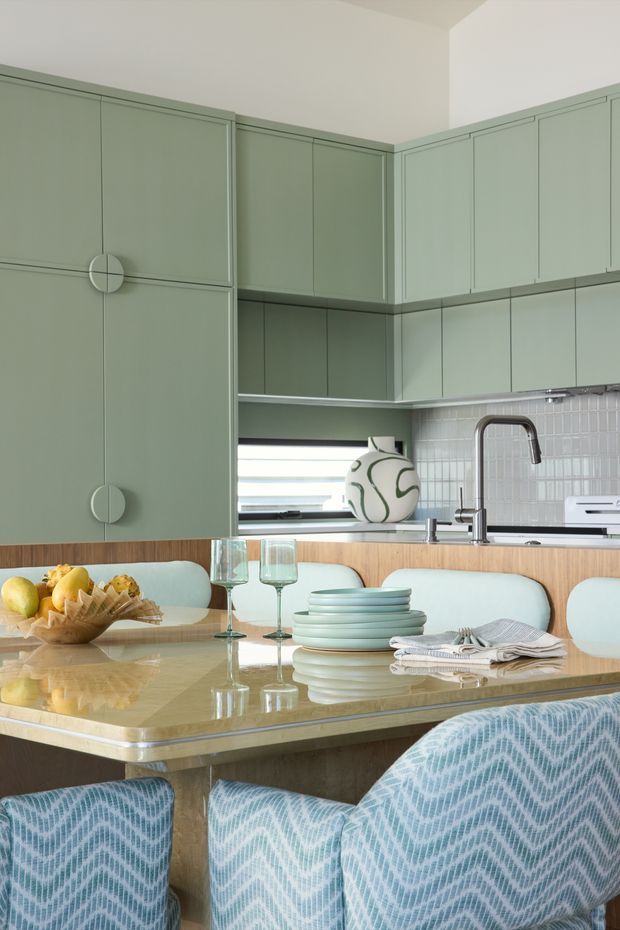 A sophisticated kitchen and dining area with muted sage-green built-in cabinetry, framed panel doors, integrated circular pulls, and a white tiled splashback. A polished pale stone island with a timber-toned base forms the dining surface, surrounded by upholstered dining chairs in a blue-and-white geometric pattern. Glassware, stacked plates, folded napkins, fruit, and a decorative vase create a styled tablescape. The space is bright, refined, and softly colourful.