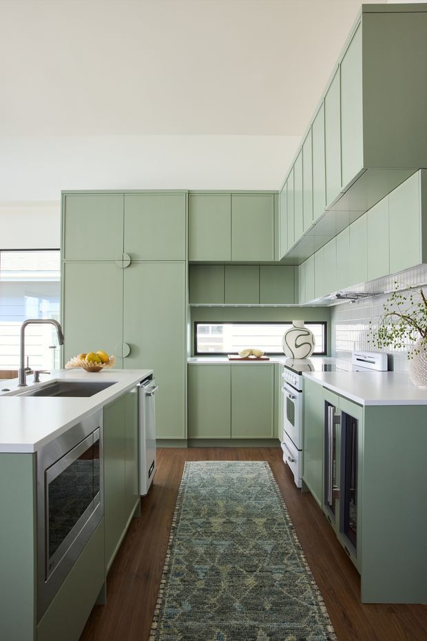 A compact, highly functional galley kitchen with pale sage-green full-height cabinetry, matching upper cabinets, white countertops, and a white tiled backsplash. Stainless-steel appliances and sink fixtures line the two opposing kitchen runs. A long patterned runner sits over dark timber flooring, while a narrow horizontal window brings in daylight. The room has a clean, tailored, retro-influenced contemporary feel.