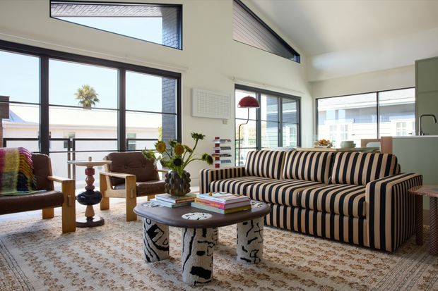 Bright contemporary living room with high ceilings, clerestory glazing, and large black-framed windows. A bold striped sofa faces a dark coffee table with sculptural patterned legs, complemented by leather lounge chairs, a patterned area rug, a red pendant lamp, books, and plants. The room opens visually toward a compact kitchen area and an adjacent outdoor terrace or balcony.