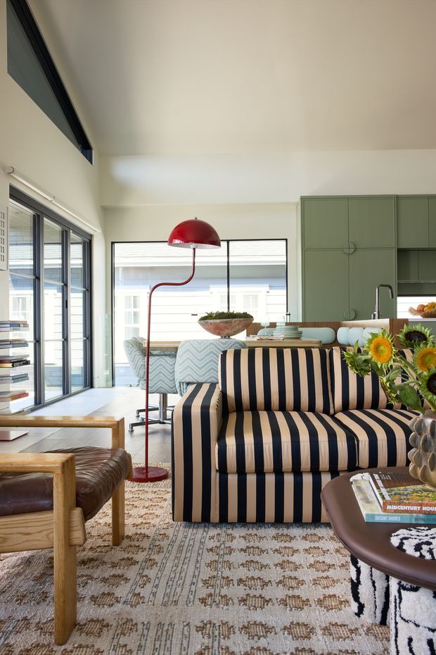 Bright, eclectic open-plan living space with a prominently striped navy-and-cream sofa, mid-century-inspired timber lounge chair, patterned rug, round coffee table, red arched floor lamp, and abundant natural light. A green built-in kitchen with panelled cabinetry is visible behind the seating area, along with large glazed openings and a dining area.