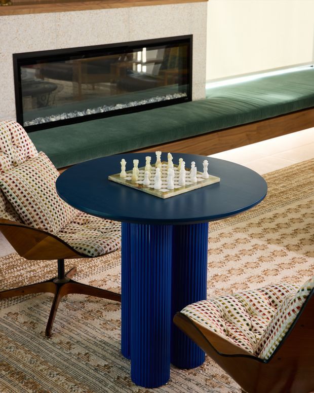 A richly styled living room seating area featuring a round deep-blue coffee table with a fluted pedestal base, a chessboard, and two patterned mid-century lounge chairs. A long dark green upholstered built-in bench runs beneath a black-framed linear fireplace set into a pale textured stone wall. Layered neutral rugs, warm timber trim, and reflective surfaces create a refined, eclectic atmosphere.