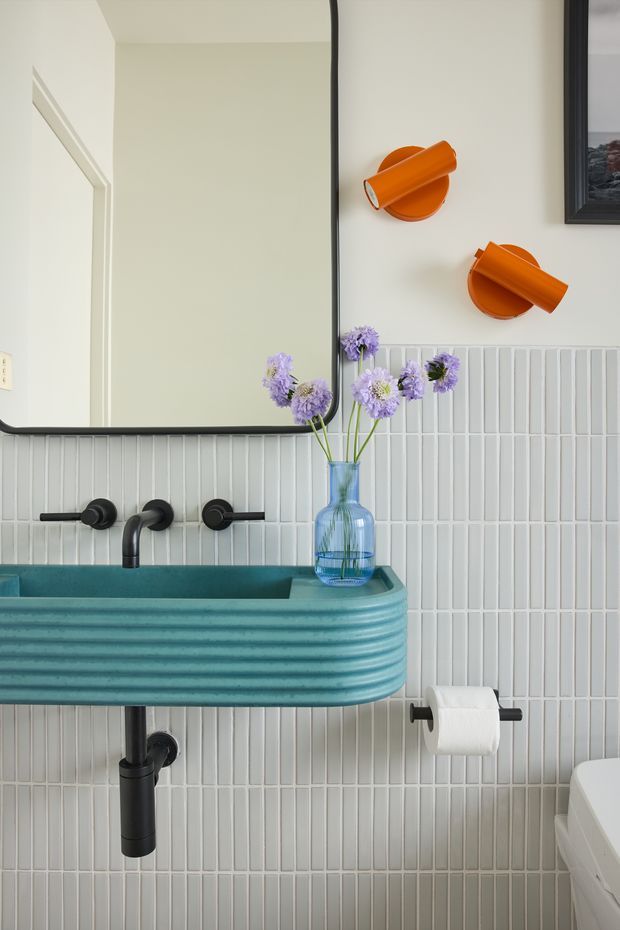 A compact, brightly styled bathroom featuring a ribbed turquoise wall-mounted vanity with integrated basin, matte black wall-mounted tapware and exposed drain, and pale vertical rectangular wall tiles. A large black-framed mirror reflects a doorway, while two orange wall sconces and a blue glass vase with purple flowers add colourful accents. A toilet is partially visible at the right.