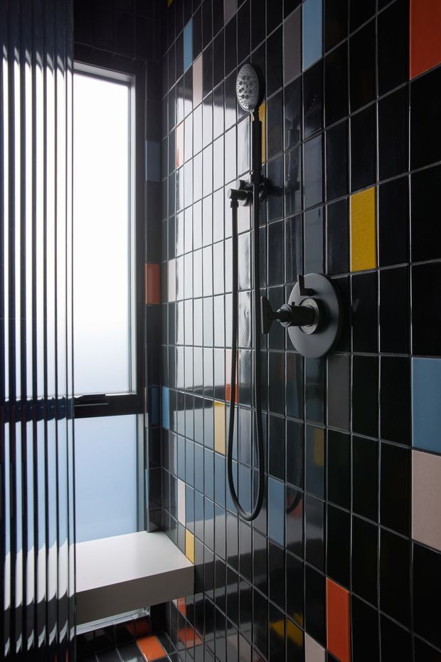 A compact contemporary shower room featuring glossy black square wall tiles with scattered geometric accent tiles in yellow, blue, red, and pale pink. A wall-mounted round rain shower and handheld shower set sit beside a large dark-framed frosted window with vertical privacy fins. The space has a bold, graphic, high-contrast aesthetic.
