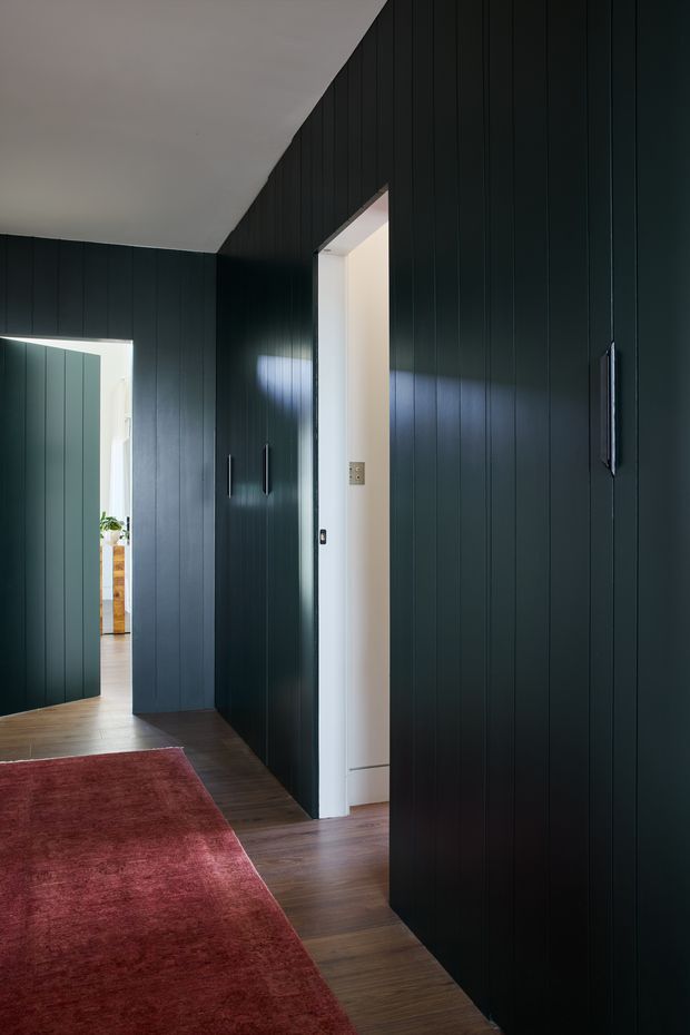 A narrow, moody interior hallway lined with dark vertical timber wall panelling and integrated dark doors or cabinetry. Warm timber flooring and a large burgundy-red rug lead toward bright adjoining rooms, while a white doorway and daylight create contrast. The space has a clean, minimalist architectural character with concealed or flush joinery.