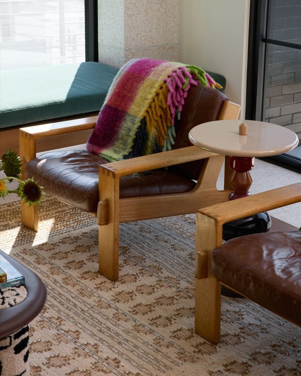 A warmly lit mid-century-inspired sitting area with a brown leather lounge chair and matching ottoman, chunky timber arms and legs, a colourful fringed wool throw, and a small round side table. A built-in window seat is visible beside large black-framed glazing, with a textured neutral rug and small indoor plants completing the relaxed interior.