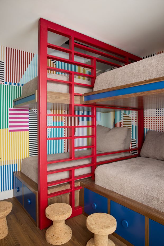 A vibrant, compact kids' bunk room with custom built-in bunk beds and seating. Bright red ladder-like framing divides the bunk structure, with blue storage cabinetry, natural timber trim and flooring, beige upholstered mattresses and cushions, and playful multicoloured geometric striped and dotted wall coverings. Two small rounded timber stools sit in the foreground. The design is highly graphic, colourful, and space-efficient.