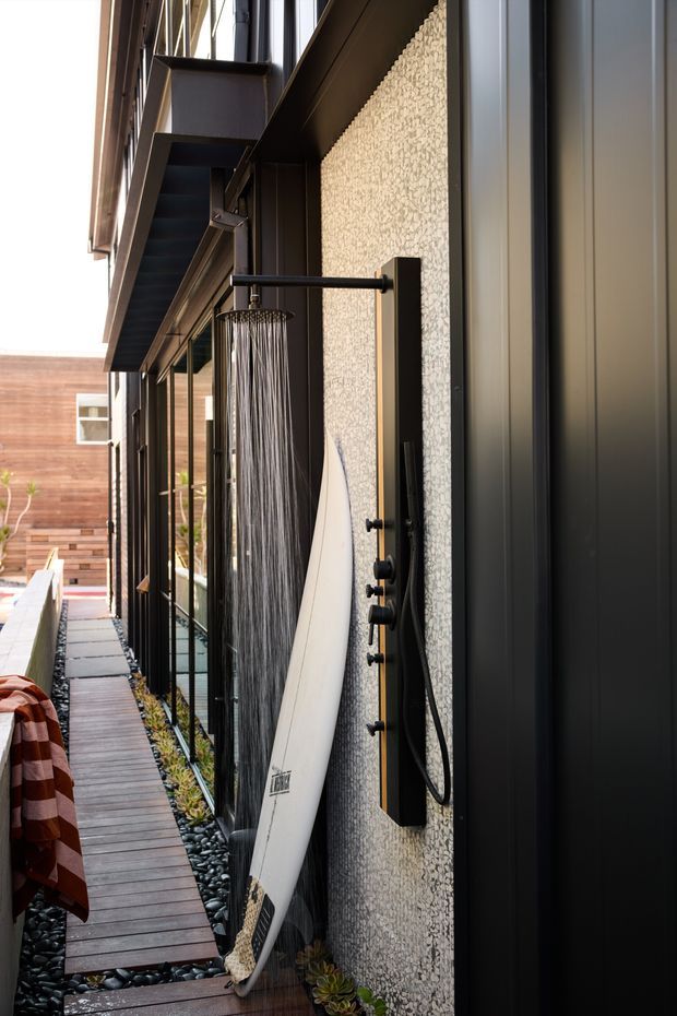 A narrow contemporary outdoor side passage beside a dark-clad building, featuring an external black shower mounted on a textured light pebble-finish wall. A surfboard leans against the wall, with a timber boardwalk, black pebbles, glass balustrade, and planted edging running through the space. Dark metal framing, balconies, and vertical cladding create a modern architectural character.