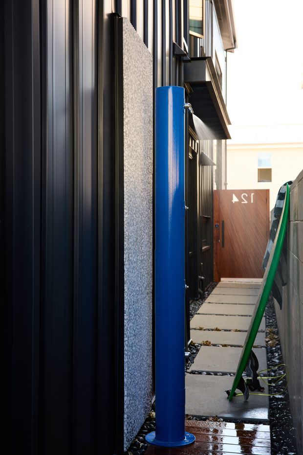 A narrow exterior side passage between buildings, featuring dark vertical metal wall cladding on the left, a textured light-coloured wall panel, a tall bright blue cylindrical outdoor fixture, and concrete stepping slabs set in dark decorative gravel. A timber-clad door and small balcony structure are visible at the far end, with a green ladder along the right wall.