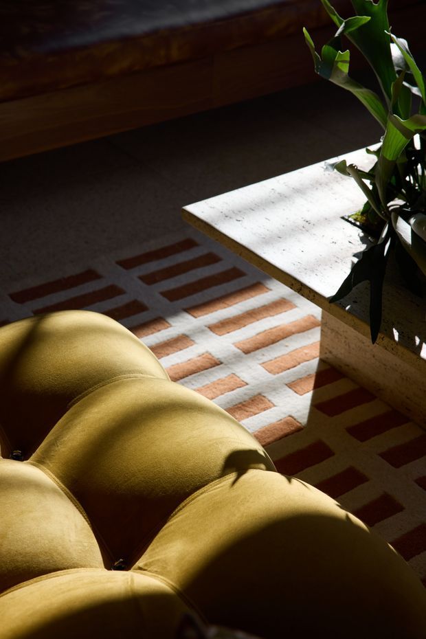 A close-up, moody interior detail showing a mustard-yellow upholstered sofa in strong sunlight, a geometric rug with cream, rust and brown rectangular blocks, a pale stone or terrazzo-topped coffee table, and a leafy indoor plant. Dark timber joinery or furniture is visible in the background, with dramatic light and shadow across the room.