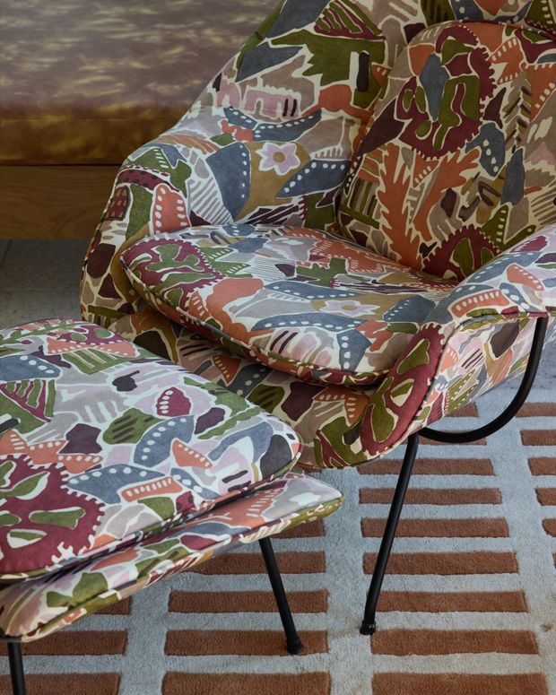 Close-up interior view of a patterned upholstered lounge chair with a matching ottoman, featuring bold multicoloured abstract botanical motifs. The furniture has slender black metal legs and sits on a geometric striped rug, with timber flooring and a muted wall visible in the background. The image emphasises colourful textile upholstery and mid-century-inspired furniture forms.