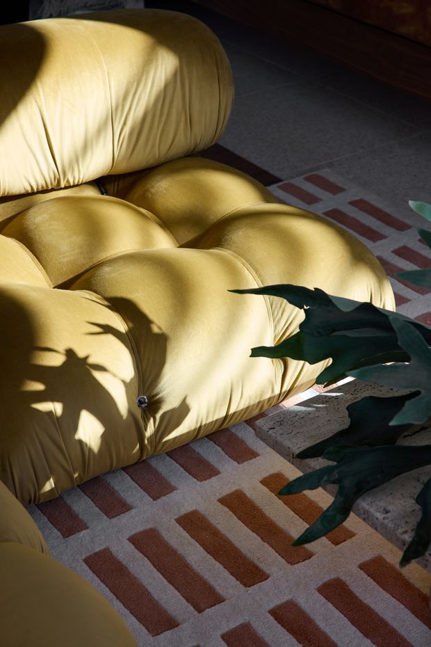 Close-up interior view of a sculptural, tufted mustard-yellow modular sofa with rounded cushions, resting on a cream rug with terracotta geometric stripes. Strong sunlight casts plant shadows across the upholstery and rug, creating a warm, tactile and dramatic composition. A dark green leafy plant partially enters the foreground.