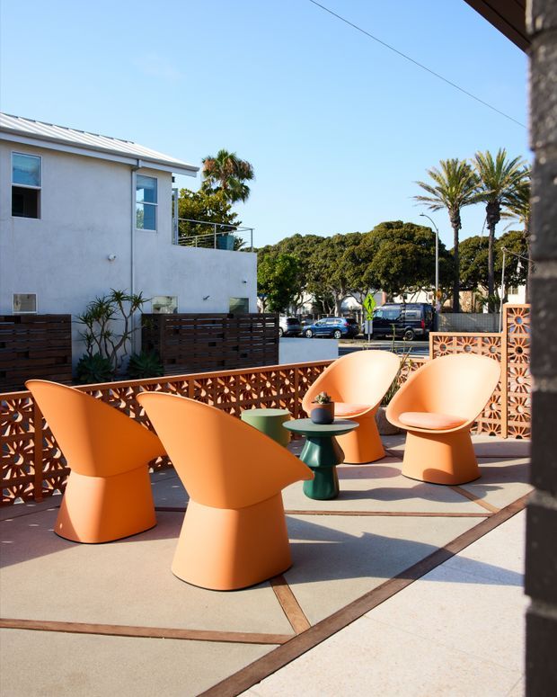 A sunny outdoor terrace furnished with several sculptural orange lounge chairs around small green side tables. The terrace has a decorative terracotta breeze-block privacy screen, pale concrete paving with dark linear inlays, and a low timber privacy fence. Beyond the terrace is a suburban street with palm trees and low-rise buildings. The scene has a distinctly mid-century-inspired, playful outdoor design.