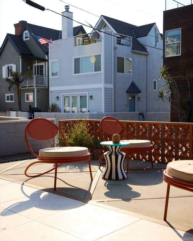 A sunny rooftop or elevated outdoor terrace furnished with sculptural retro-style orange lounge chairs, round cream cushions, and a small striped pedestal side table. A perforated terracotta-toned privacy screen, pale concrete paving, and potted plants define the terrace, with tightly packed multi-storey residential homes visible beyond.