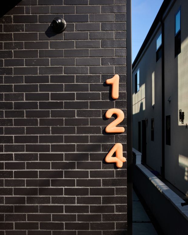 Close-up architectural exterior view of a dark charcoal brick wall with raised orange address numbers 1, 2, and 4 and a black wall-mounted light fixture. A narrow side passage and the facade of an adjacent modern building are visible to the right, strongly lit by low-angle sunlight and contrasted against blue sky.