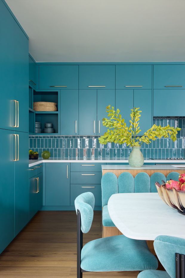 Vibrant teal-blue kitchen with full-height built-in cabinetry, brass handles, open shelving, and a glossy blue-and-white tiled splashback. A peninsula or island separates the kitchen from a dining area with a rounded white table and upholstered turquoise dining chairs. Dark timber flooring and a pale ceiling balance the saturated cabinetry, while a vase of foliage adds a natural accent.