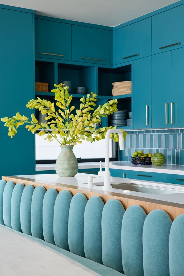 A brightly styled kitchen dominated by teal-blue built-in cabinetry with brass-toned handles. The space features open shelving, a white countertop and sink with a sculptural white tap, and a blue-green tiled splashback. In the foreground, a large island has a light timber edge and a distinctive upholstered scalloped bench in pale blue. A leafy vase and fruit bowl add organic colour and texture.