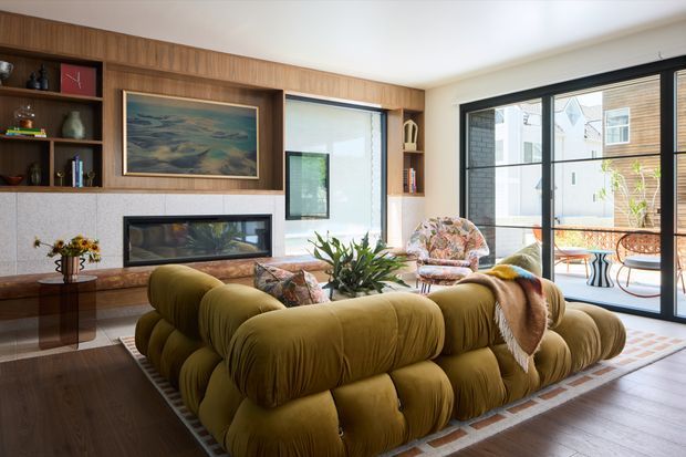 A bright, richly layered living room with a large olive-green modular sofa arranged around a low coffee table. A timber built-in wall combines open shelving, cabinetry and a recessed fireplace with a pale stone or tiled surround. Large black-framed glazed doors and windows open to a small outdoor terrace or courtyard. Decorative artwork, books, plants, patterned cushions and a floral accent chair add warmth and texture. The room has dark timber flooring, a light ceiling and a refined mid-century-inspired contemporary character.