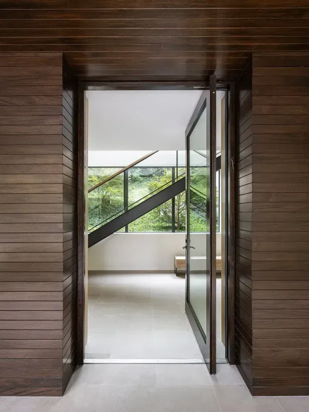 A contemporary interior view through a tall open glazed doorway into a bright landing or circulation space. Dark horizontal timber wall and ceiling cladding frame the opening, while a black steel-and-glass stair balustrade and diagonal stair structure are visible beyond. Pale tiled flooring, white walls, and abundant glazing create a clean, modern connection to lush greenery outside.