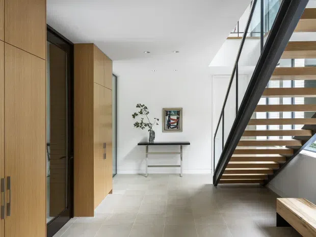 Bright contemporary entry hall with a floating open-riser staircase, black metal and glass balustrade, extensive warm timber built-in cabinetry, pale tiled flooring, and a minimalist console table. A skylight or high-level glazing brings natural light into the circulation space.