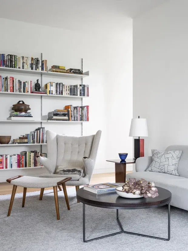 Bright, minimalist living room with built-in open shelving filled with books and objects, a tufted wingback lounge chair, matching ottoman, pale sofa, round dark coffee table, and a floor lamp. The room has white walls, light grey carpet, and a restrained mid-century-inspired furnishing palette.