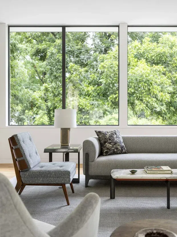 A bright, refined living room with expansive black-framed windows overlooking dense greenery. The room features a grey upholstered sofa, a patterned accent cushion, a matching textured lounge chair with timber arms and legs, a small glass-and-metal side table with a table lamp, and a low marble-topped coffee table. Neutral grey and white finishes create a calm, contemporary interior.