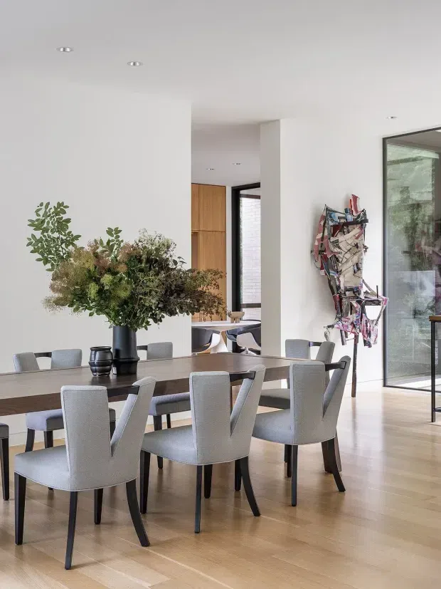 Bright contemporary dining area with a long timber dining table and upholstered grey dining chairs. White walls and ceiling, pale timber flooring, recessed downlights, black-framed floor-to-ceiling glazing, and a timber-lined opening toward the adjoining space create a clean, refined interior. A large floral arrangement and a sculptural wall artwork provide decorative accents.