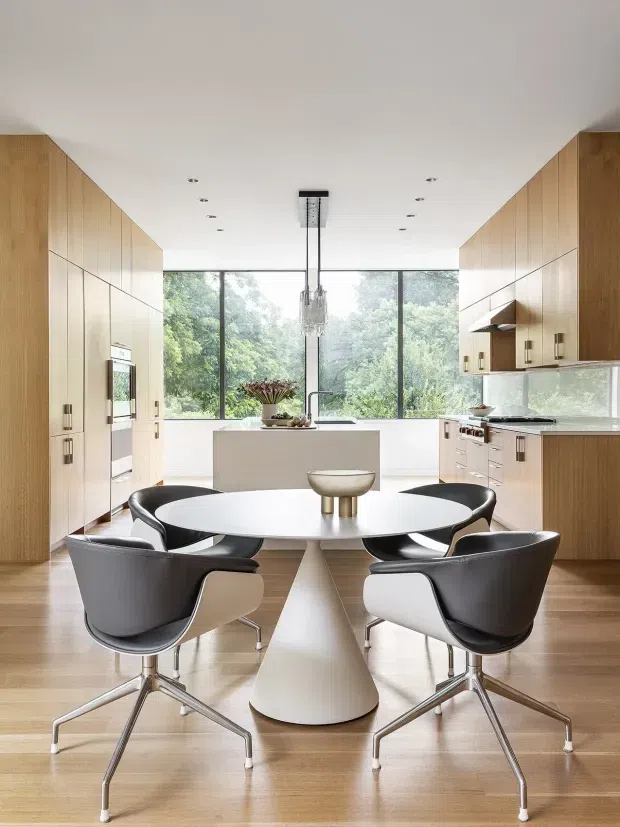 A bright, refined open-plan kitchen and dining space. Full-height light timber cabinetry lines both sides of the room, with integrated appliances and pale benchtops. A central island sits in front of expansive floor-to-ceiling glazing overlooking leafy trees. In the foreground, a round white dining table with sculptural pedestal base is surrounded by four black and white contemporary dining chairs. A crystal pendant hangs above the island, and warm timber flooring runs throughout.