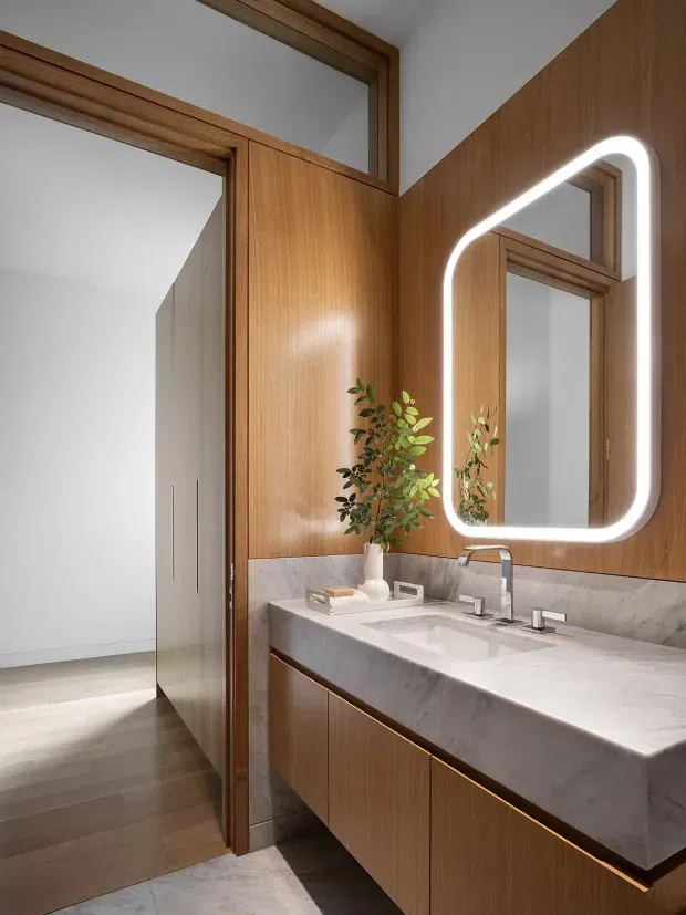A refined contemporary bathroom vanity with a long double-sink basin, substantial pale marble or engineered-stone benchtop, timber-veneer cabinetry, matching timber wall panelling, and a large rounded-rectangle illuminated mirror. Timber-framed glazing or internal openings connect visually to a bright adjacent space. The room has a warm, understated palette and clean custom joinery.
