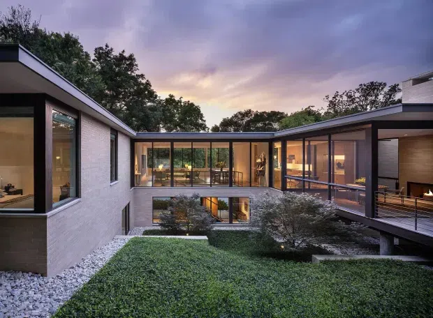 Twilight exterior view of a contemporary multi-wing residence arranged around a landscaped internal courtyard. The home features flat rooflines, pale masonry walls, extensive floor-to-ceiling glazing, dark slender window frames, timber-lined interiors, and a raised terrace or bridge-like connection overlooking the garden.