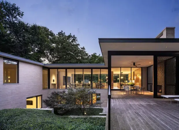 Contemporary low-profile residence arranged around a landscaped courtyard, photographed from an elevated outdoor deck at dusk. The home features broad floor-to-ceiling glazing, pale brick walls, dark slim metal framing, flat roof forms, warm interior lighting, and a timber deck extending toward an open-plan living and dining area.
