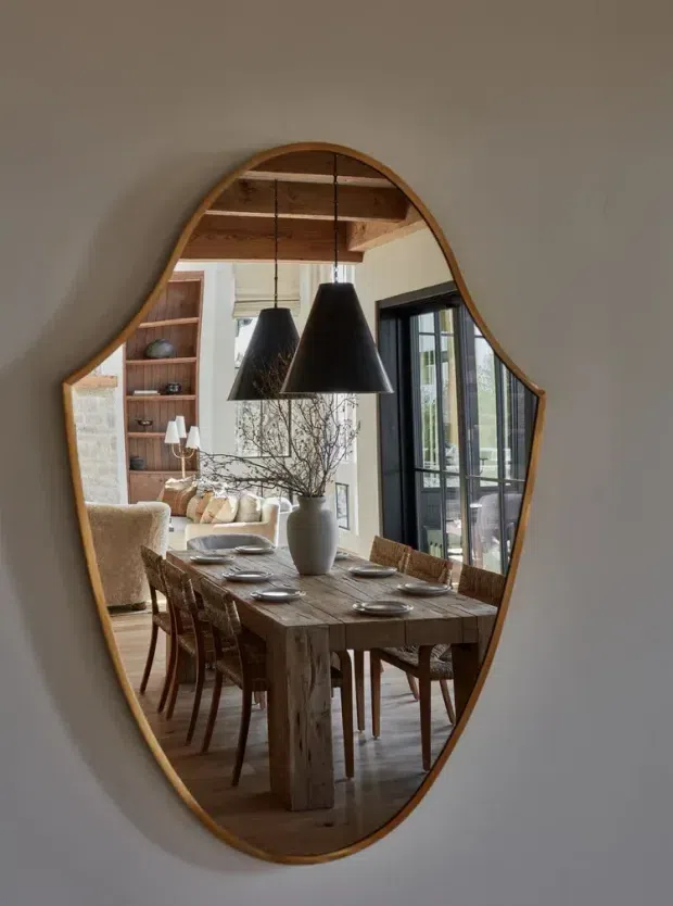 A vertically oriented organic-shaped wall mirror reflects a warm dining area with a substantial rustic timber dining table, woven dining chairs, dark pendant lights, timber ceiling beams, and a bright adjoining living space. Black-framed glazed doors and shelving are visible in the reflection, creating a relaxed, natural, contemporary-rustic interior.