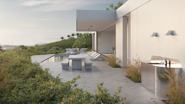 A contemporary flat-roofed house with a minimalist pale facade and expansive outdoor terrace overlooking a landscaped hillside. The terrace includes a reflecting pool, concrete dining table and benches, loungers, and a compact stainless-steel outdoor kitchen with integrated sink and tap. Recessed lighting, clean geometric volumes, and sparse ornamental grasses reinforce the refined modern aesthetic.