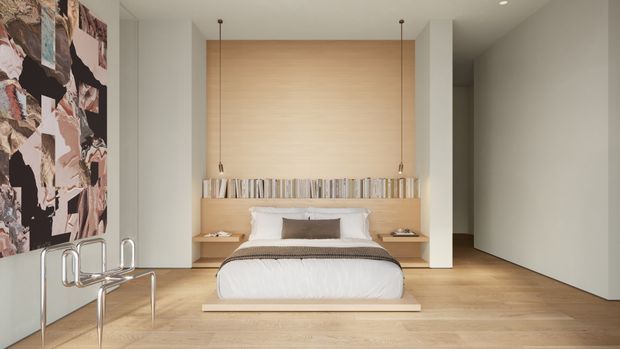 A minimalist bedroom with a low platform bed centered against a full-height light timber feature wall. Integrated bedside surfaces, narrow pendant lights, pale timber flooring, and a large abstract artwork create a restrained contemporary interior. A sculptural chrome chair sits in the foreground, while a concealed doorway opens to the right.