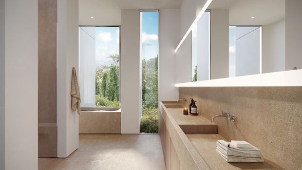 Bright contemporary bathroom with a long integrated stone vanity, wall-mounted basin and tapware, large frameless mirrors, warm beige textured wall and floor finishes, and tall windows providing garden views. A freestanding bathtub is visible at the rear, with minimalist open shelving and concealed lighting contributing to the clean, refined atmosphere.