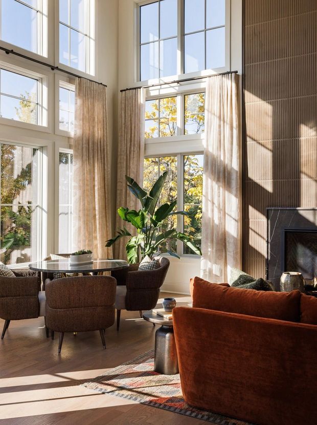 Sunlit double-height living room with extensive multi-pane floor-to-ceiling glazing, tall sheer curtains, a sculptural stone fireplace, a round glass dining table with upholstered chairs, a rust velvet sofa, indoor greenery, and warm timber flooring. Vertical ribbed wall panelling adds texture beside the fireplace.
