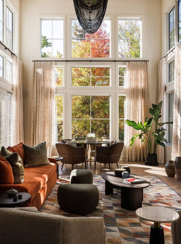 A bright, double-height living room with expansive multi-pane windows, full-height sheer curtains, and autumn trees visible outside. The room features a warm rust-orange sofa, dark upholstered lounge chairs, layered seating, sculptural black pendant lighting, a patterned geometric rug, dark oval coffee table, indoor plants, and small side tables. The overall atmosphere is refined, warm, and contemporary with strong natural light.