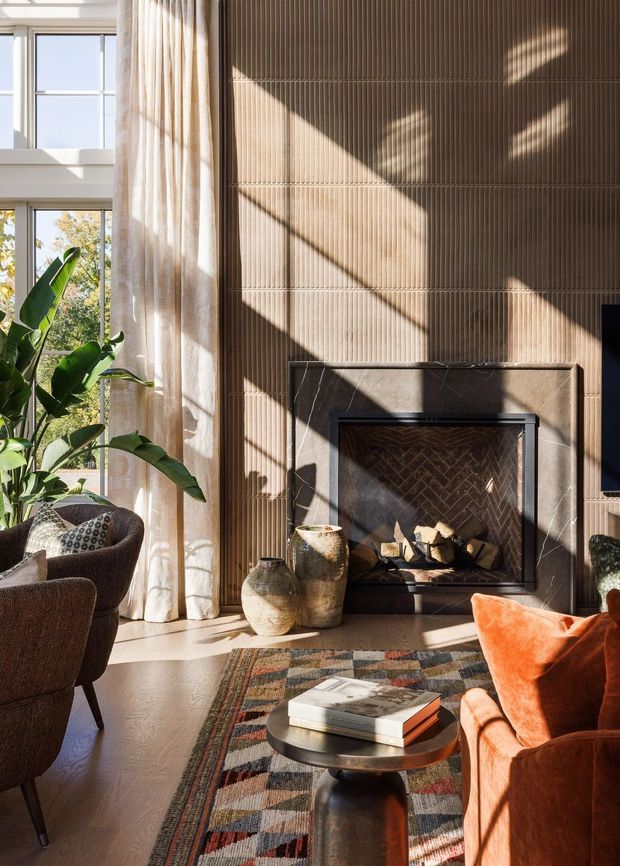 A warm, sunlit living room centered on a fireplace with a vertically ribbed beige-brown feature wall and dark stone surround. Tall multi-pane windows with sheer curtains bring in strong natural light, while upholstered lounge chairs, an orange sofa, patterned rug, round coffee table, and ceramic vessels create a layered, inviting interior.