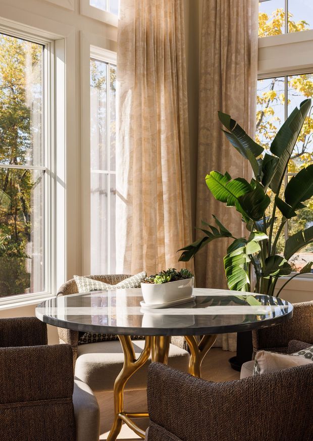 A bright, elegant dining nook surrounded by tall windows, with warm sunlight filtering through floor-to-ceiling beige curtains. A round marble-topped dining table with a sculptural brass-gold base is surrounded by textured upholstered dining chairs. A large tropical indoor plant and a small tabletop planter add organic greenery, while the windows overlook leafy trees.